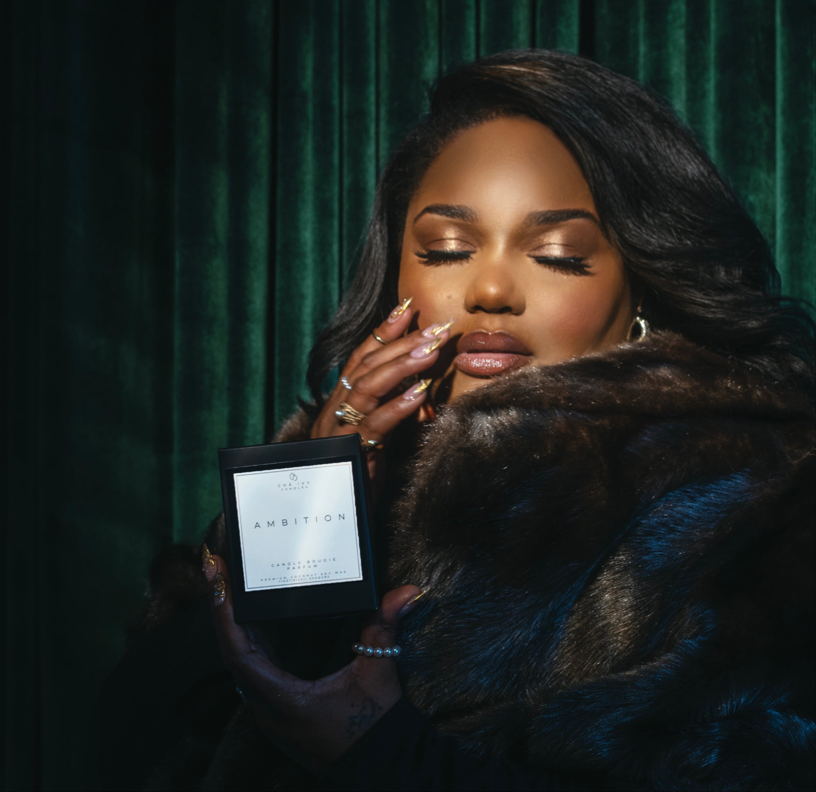 Woman holding a black box with 'Ambition' text on black brand candle box against a green curtain background